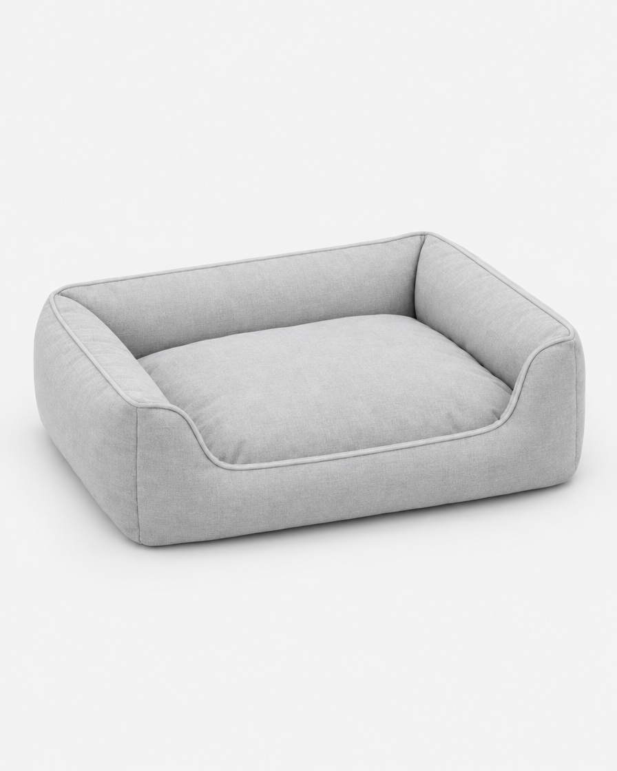 A low rectangular dog bed with a rolled bolster on three sides, in a very light cool grey woven fabric, photographed on a plain light grey studio background.