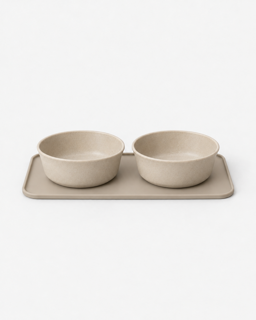 Two warm beige stoneware pet bowls sitting side by side on a sand-coloured silicone mat against a light grey studio background.
