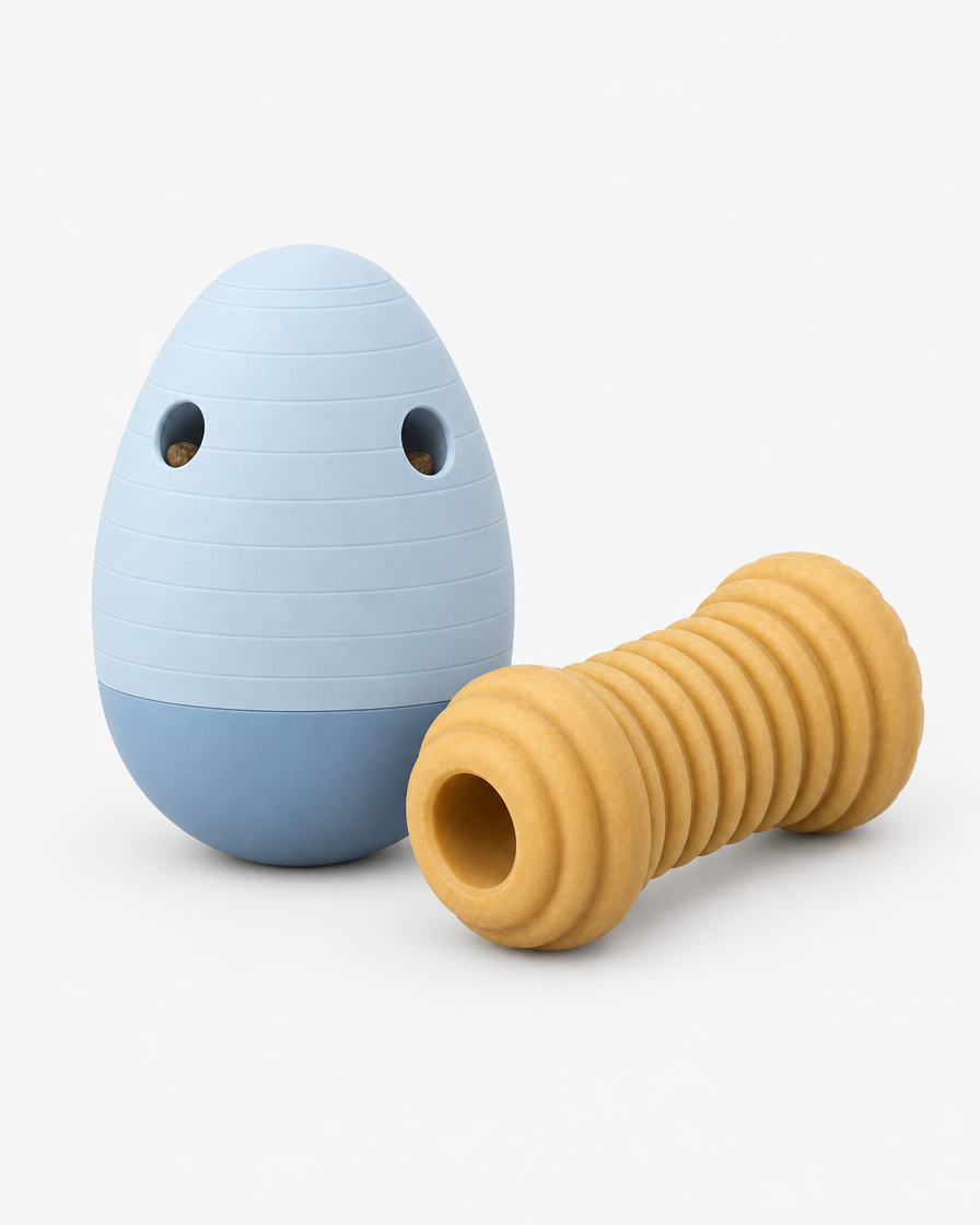 A pale blue egg-shaped dog puzzle feeder standing beside a ridged honey-coloured rubber chew toy on a light grey studio background.