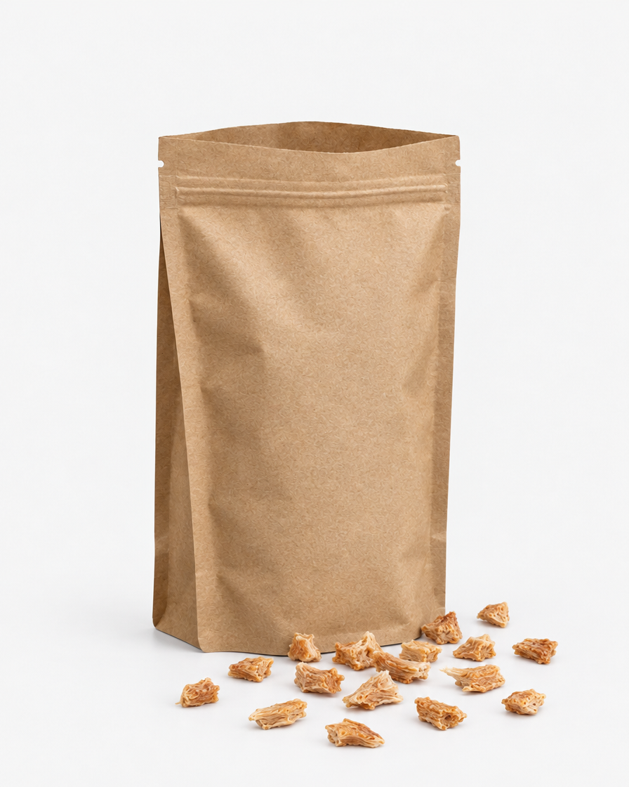 An open kraft-paper pouch with small dried chicken treat pieces spilled out beside it on a light grey background.